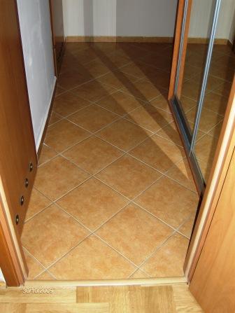 Beżowe, kwadratowe płytki podłogowe ułożone pod kątem 45 stopni w korytarzu, widok z góry, z fragmentem drewnianej listwy przypodłogowej i częścią szafy wnękowej z lustrzanymi drzwiami.