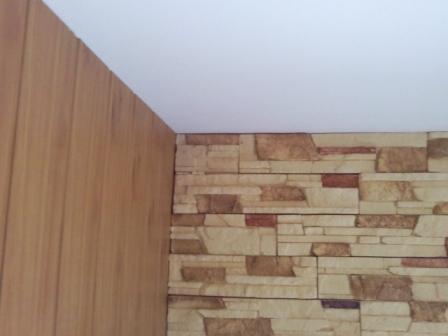 Wykończenie narożnika pomieszczenia: ściana z paneli imitujących drewno sąsiaduje ze ścianą wyłożoną płytkami imitującymi kamień. Widoczny biały sufit.