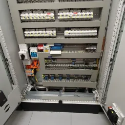 Otwarta szafa elektryczna z licznymi bezpiecznikami, przekaźnikami i okablowaniem, widok z góry na szarą podłogę z płytek.