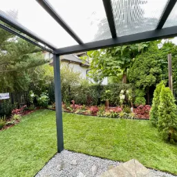 Zadbany trawnik pod zadaszeniem tarasowym, obrzeża z kamyków, widoczne nasadzenia krzewów i drzew, czarne metalowe ogrodzenie.