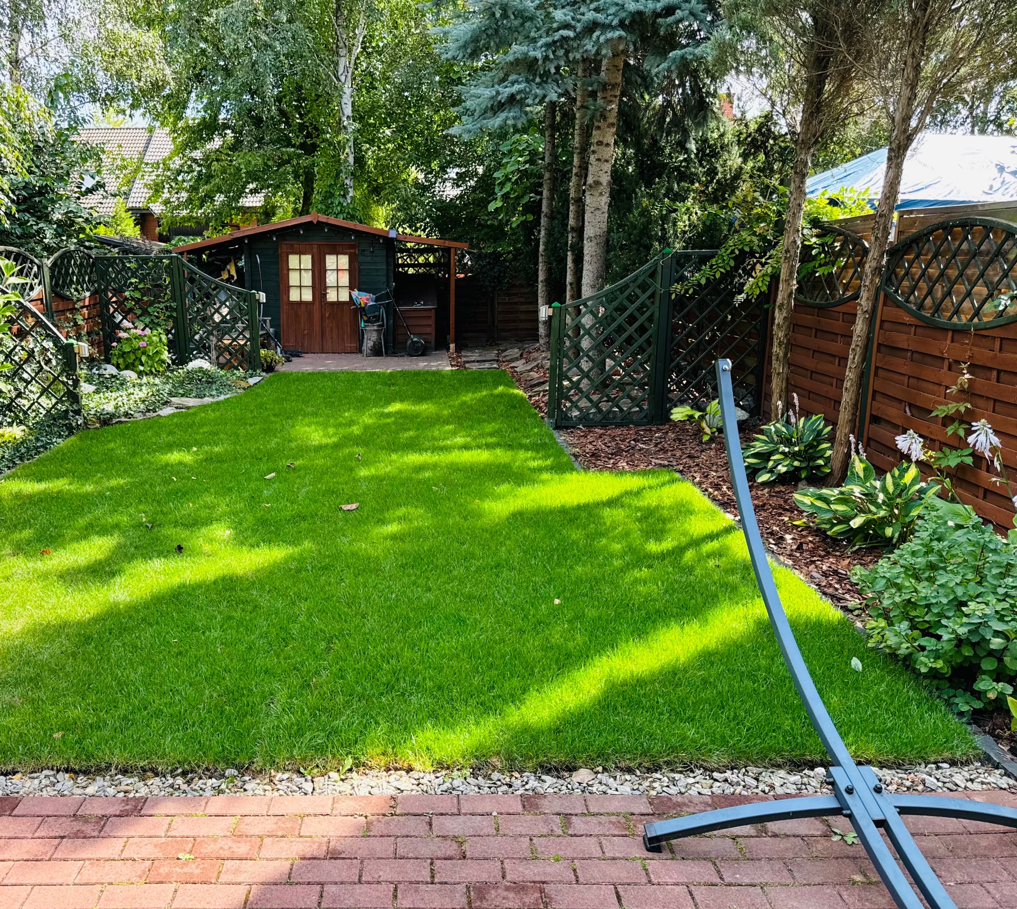 Wypielęgnowany trawnik z drewnianą altaną w tle, obrzeże z kostki brukowej i fragment stelaża do hamaka.