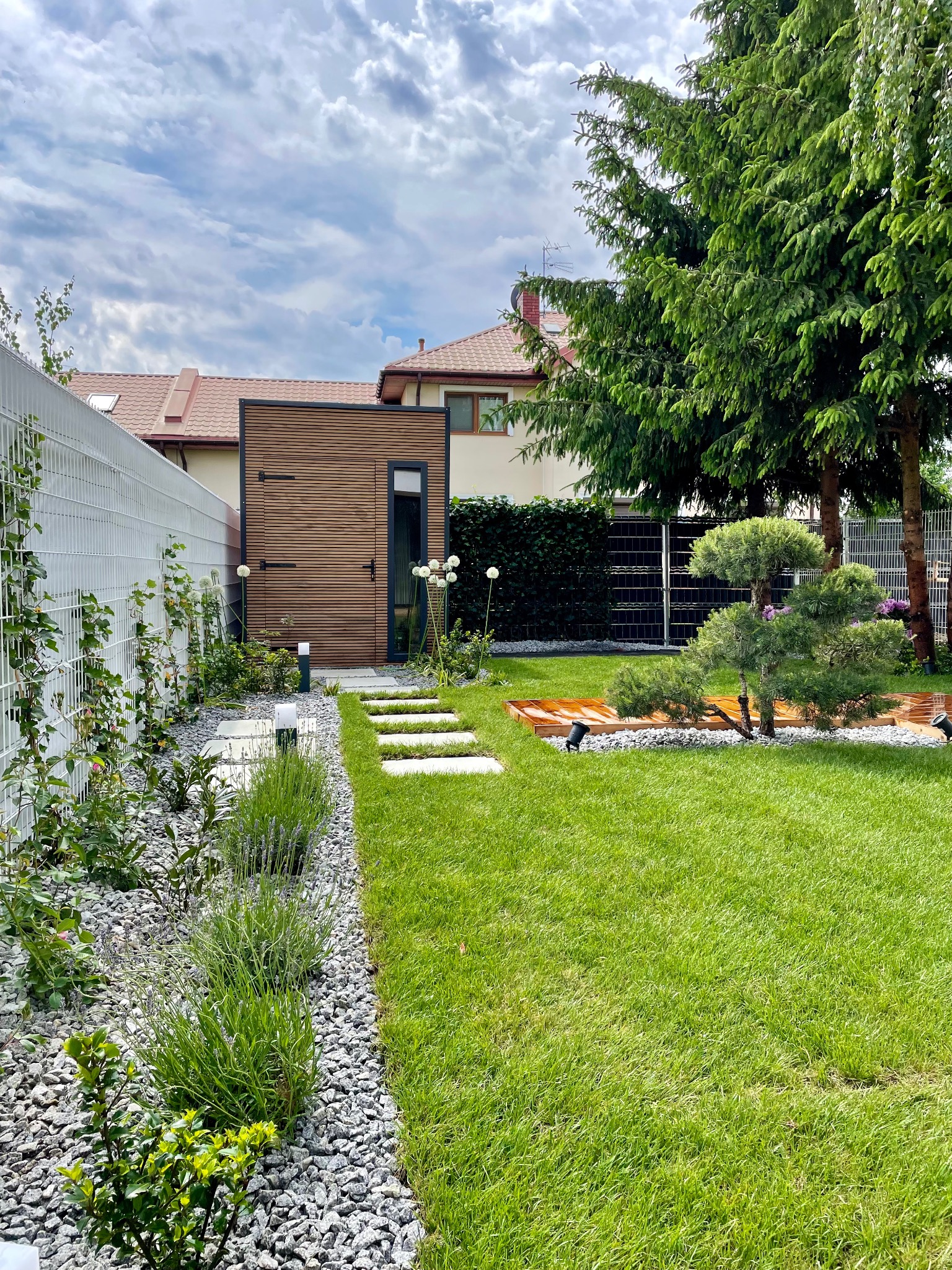 Nowoczesny ogród z równo przystrzyżonym trawnikiem, ścieżką z płyt betonowych, ozdobnymi kamieniami, roślinami, drewnianym domkiem narzędziowym i oświetleniem.