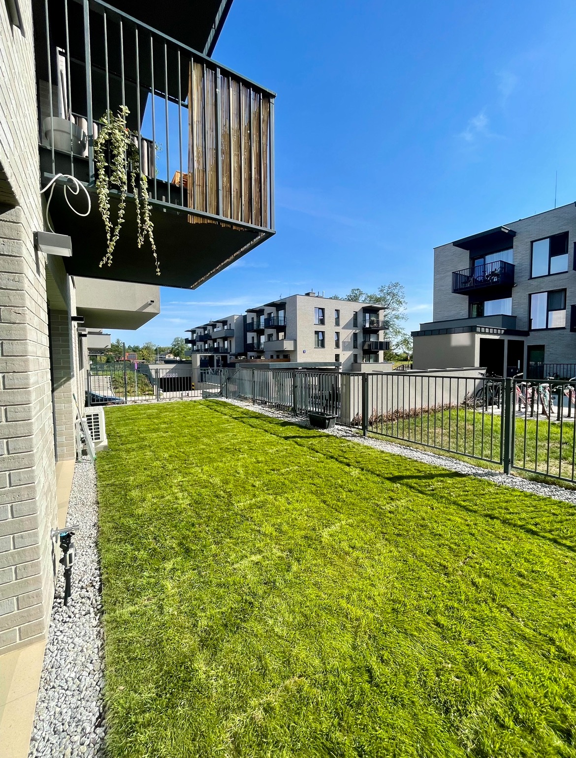 Świeżo założony trawnik przed nowoczesnym budynkiem mieszkalnym z szarej cegły, widok z poziomu gruntu, metalowe ogrodzenie oddzielające trawnik od przestrzeni wspólnej.