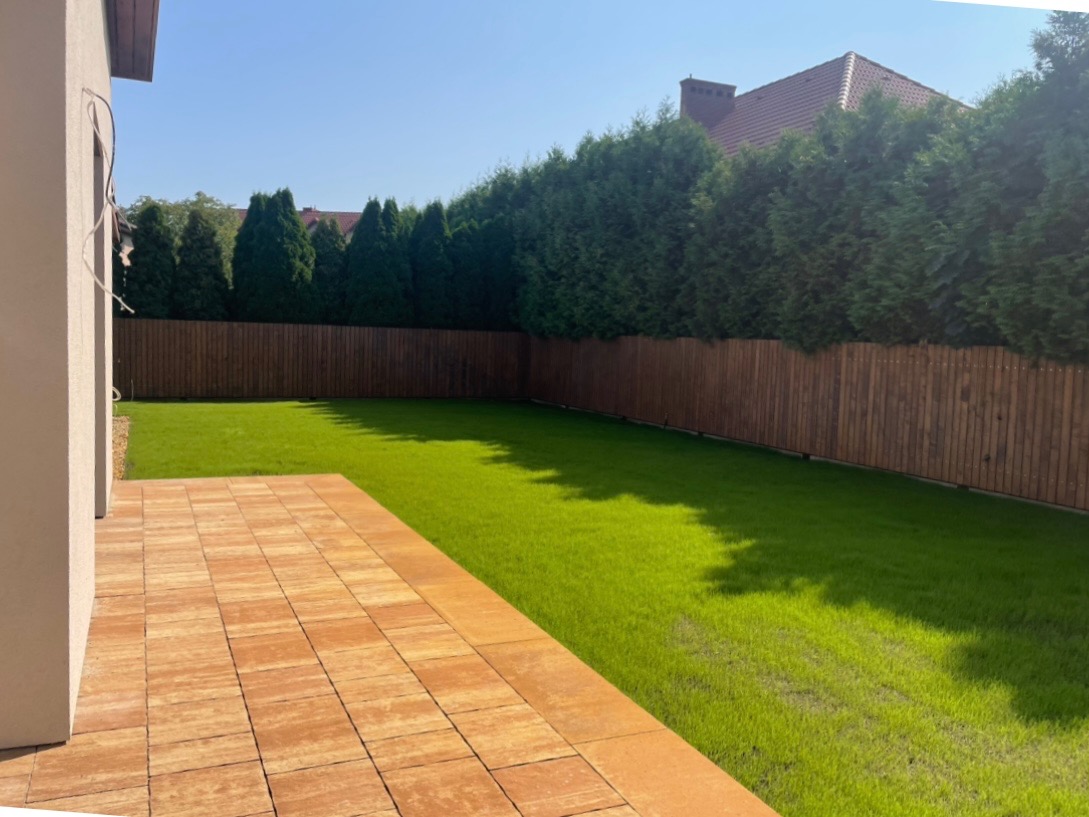 Ułożony taras z kostki brukowej w odcieniach brązu przylegający do domu, za nim trawnik i drewniany płot z nasadzeniami z iglaków.