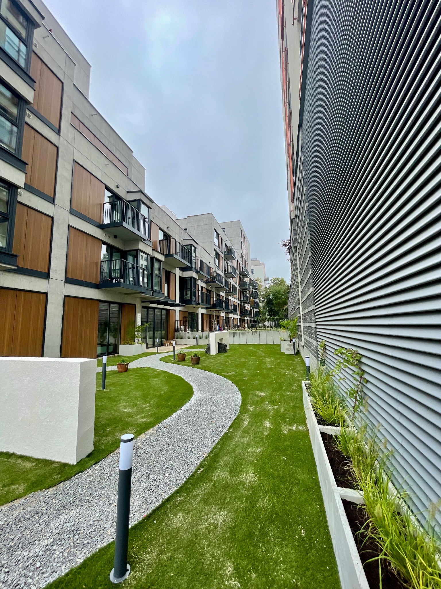 Kaskadowe budynki mieszkalne z balkonami, ścieżka z drobnego żwiru wijąca się przez trawnik, oświetlenie ścieżki i donice z roślinami ozdobnymi.