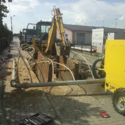 Żółta koparka w wykopie zabezpieczonym metalowymi szalunkami, rury, pompa odwadniająca Varisco, narzędzia na kostce brukowej. Prace ziemne przyłącza wod-kan.