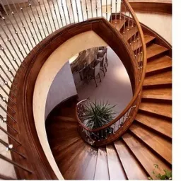 Spiralne schody drewniane z metalową balustradą, widok z góry, z doniczkową rośliną na podeście i widocznym fragmentem stołu w tle.