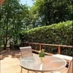 Jasny, drewniany taras z metalowym stołem i krzesłami. W tle widoczne zielone krzewy i drzewa.