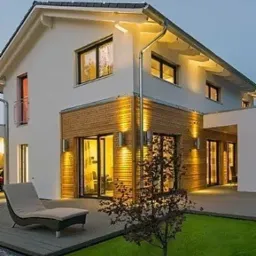 Nowoczesny dom jednorodzinny z elewacją w kolorze białym i drewnianą okładziną, oświetlony ciepłym światłem lamp elewacyjnych, z tarasem i leżakiem na trawniku.