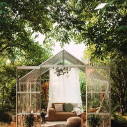 Ustawiona w ogrodzie szklarnia z otwartymi drzwiami, wewnątrz urządzona strefa relaksu z sofą, poduszkami, dywanem i roślinami doniczkowymi, całość oświetlona naturalnym światłem.