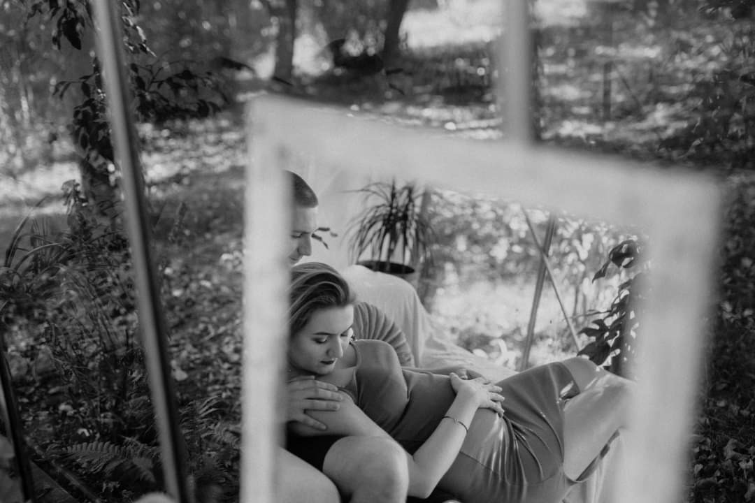 Czarno-białe ujęcie pary w objęciach, widoczne w odbiciu lustrzanym na tle leśnej scenerii z paprociami i drzewami.