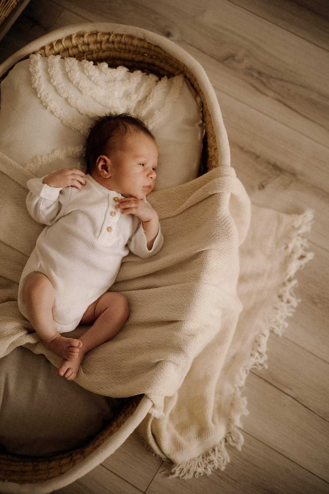 Noworodek w białym body leży w wiklinowym koszu na kremowym kocu z frędzlami, z poduszką z falbankami w tle, na podłodze z paneli.