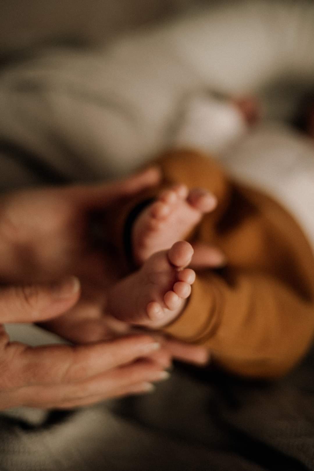 Drobne stópki noworodka w brązowych spodniach, delikatnie trzymane w dłoniach rodzica, na tle miękkiej, szarej tkaniny.