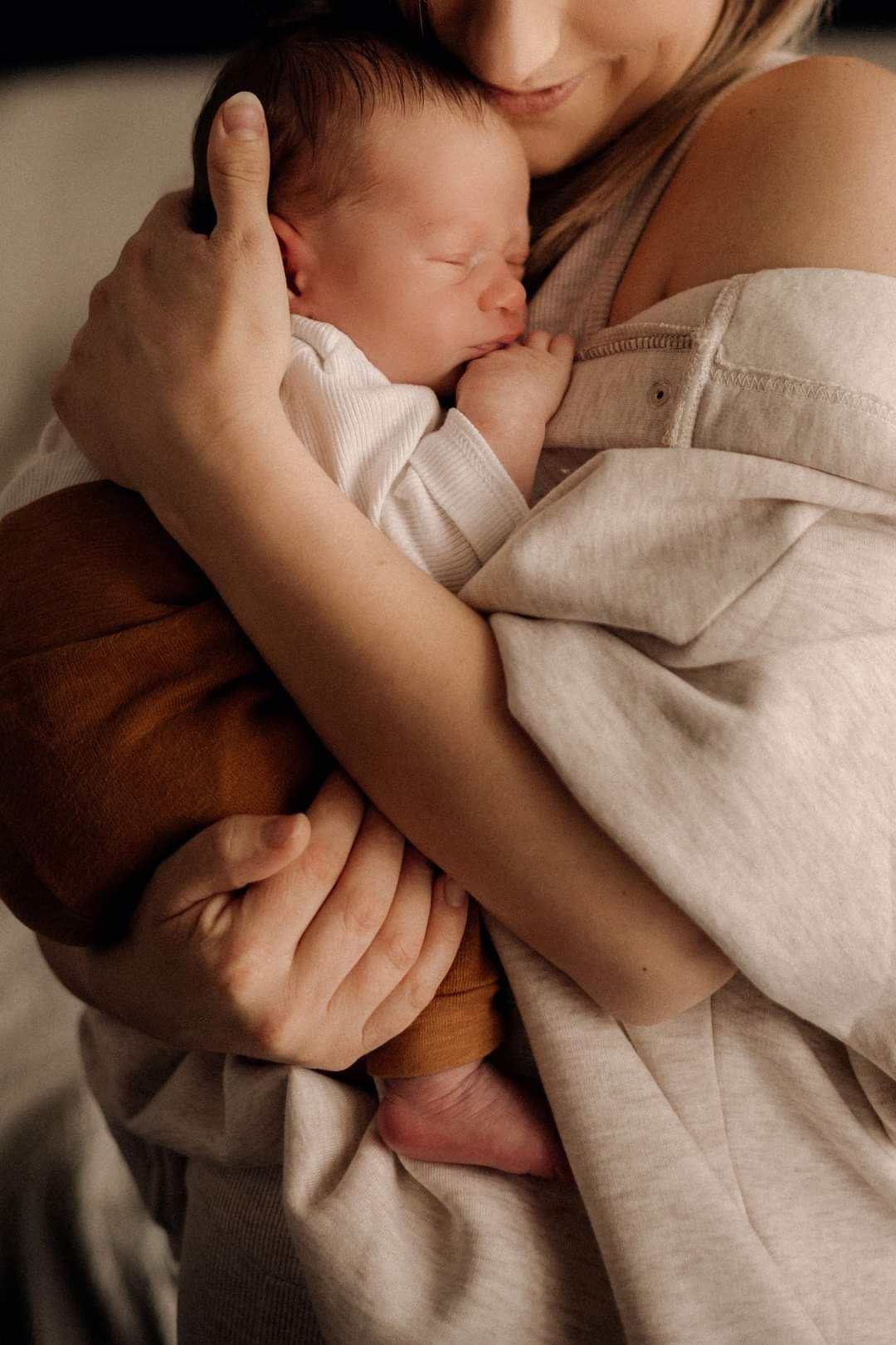 Noworodek w brązowych spodniach śpi w ramionach matki, delikatne światło podkreśla gładką skórę dziecka.