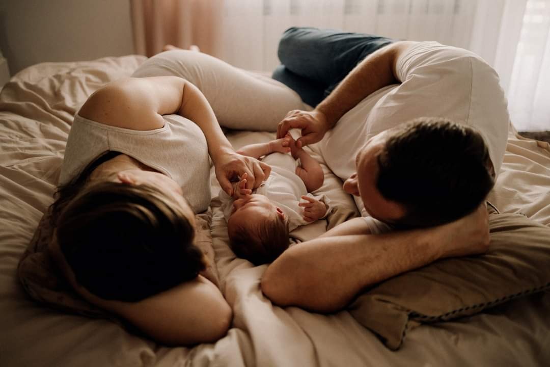 Rodzice leżący na łóżku twarzą do noworodka, delikatnie trzymający jego dłonie i stopy, w naturalnym świetle.