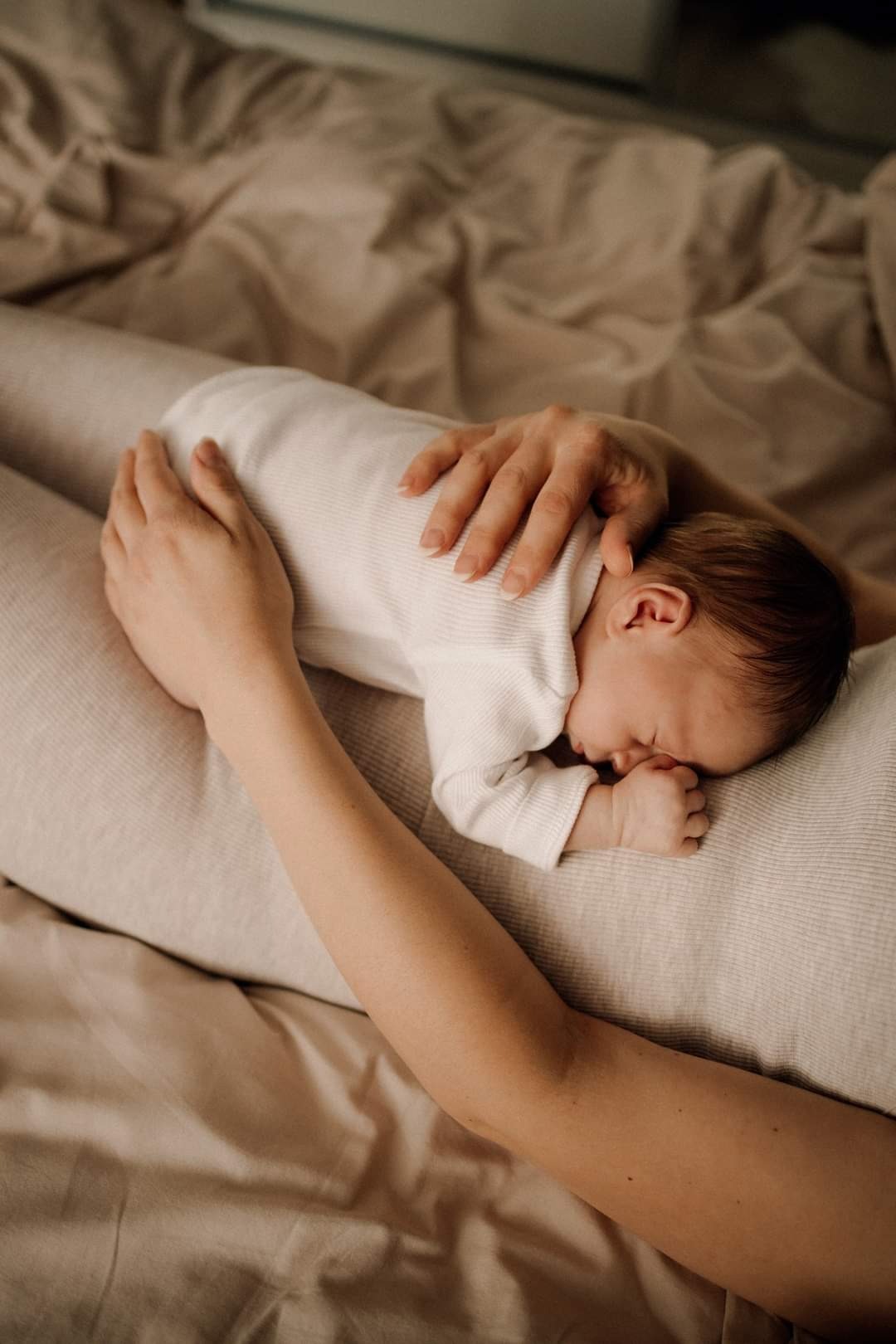 Śpiący noworodek w białym body leży na beżowej poduszce, trzymany przez dłonie matki, z widoczną drobną rączką przy twarzy.