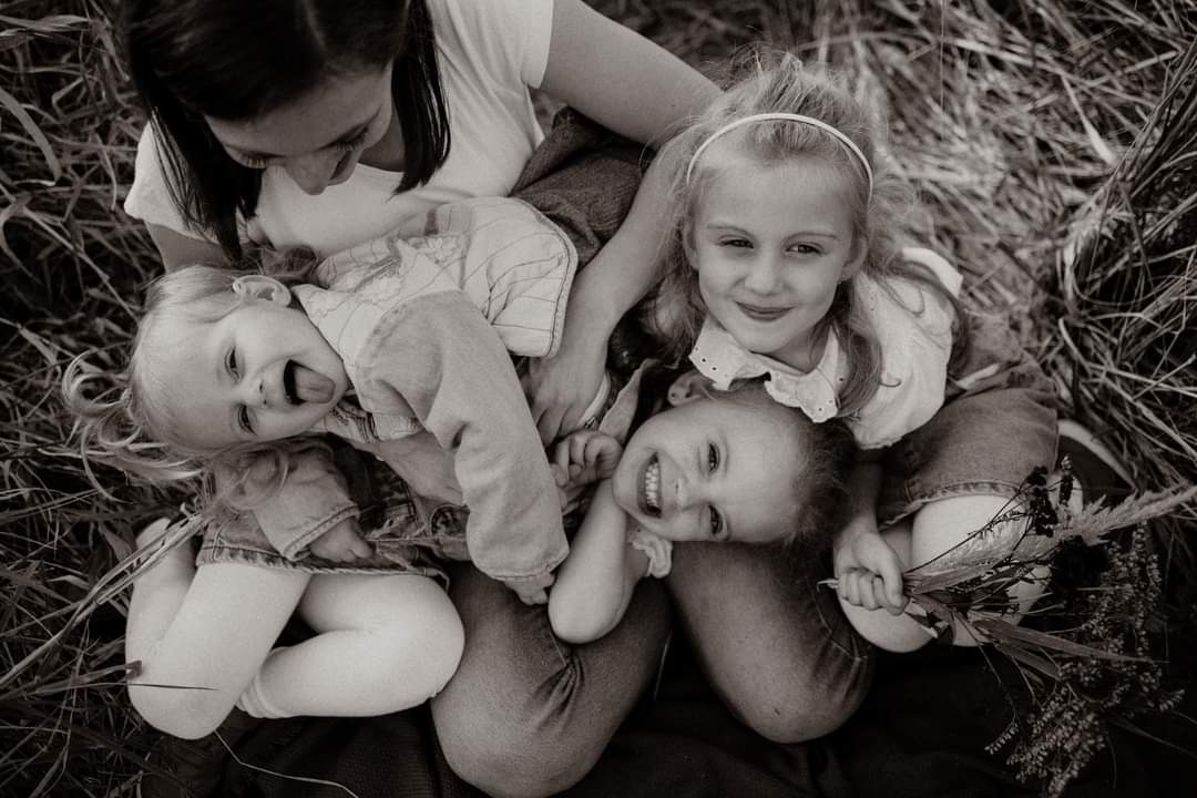 Czarno-białe zdjęcie matki z trzema córkami leżącymi na trawie, uśmiechnięte dziewczynki trzymają mały bukiet polnych kwiatów, jedna z córek pokazuje język.