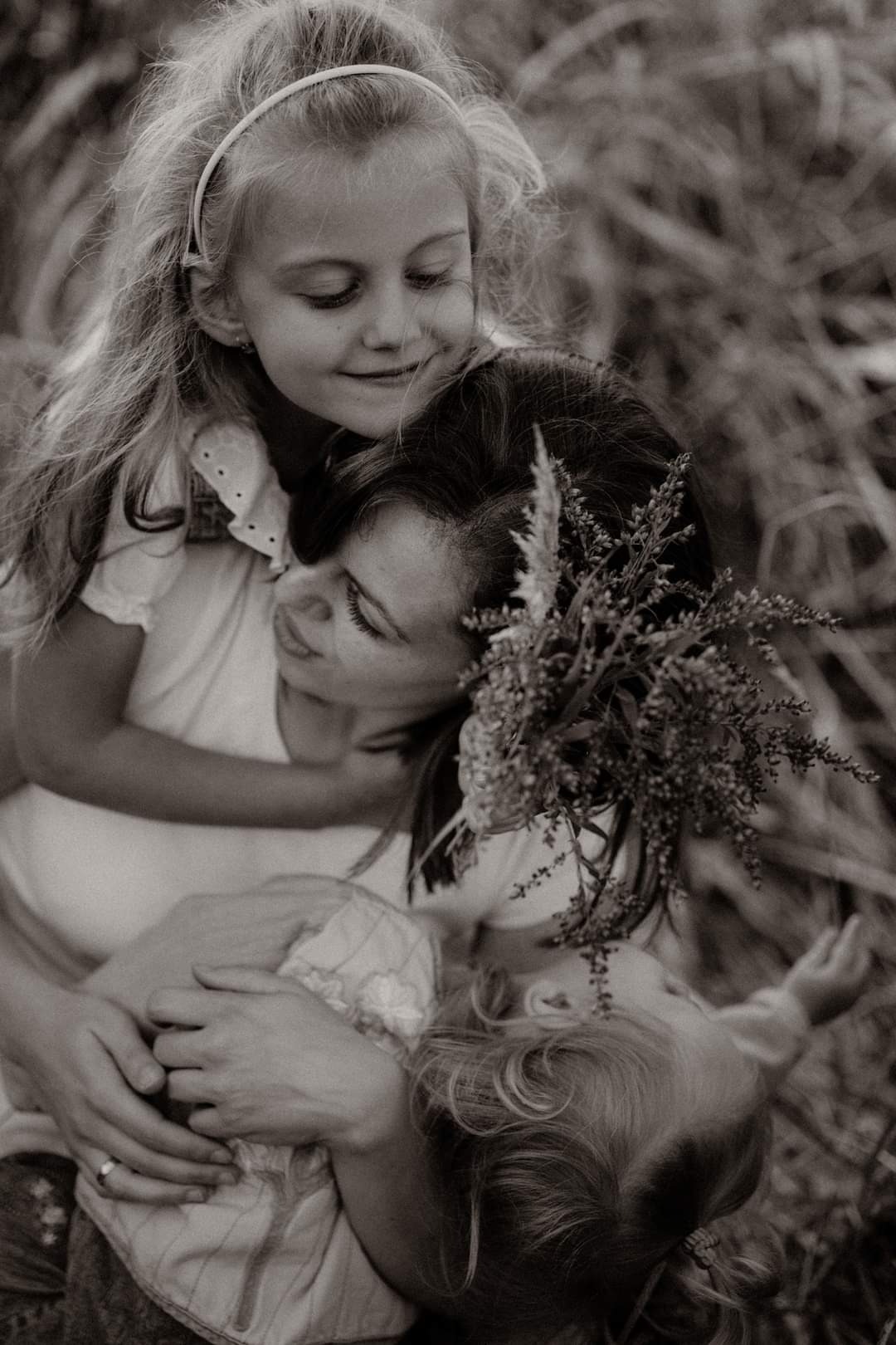 Czarno-białe zdjęcie matki obejmującej dwie córki w wysokiej trawie; starsza córka z opaską na głowie przytula się do matki, młodsza leży w jej ramionach, matka ma we włosach wpięte polne kwiaty.