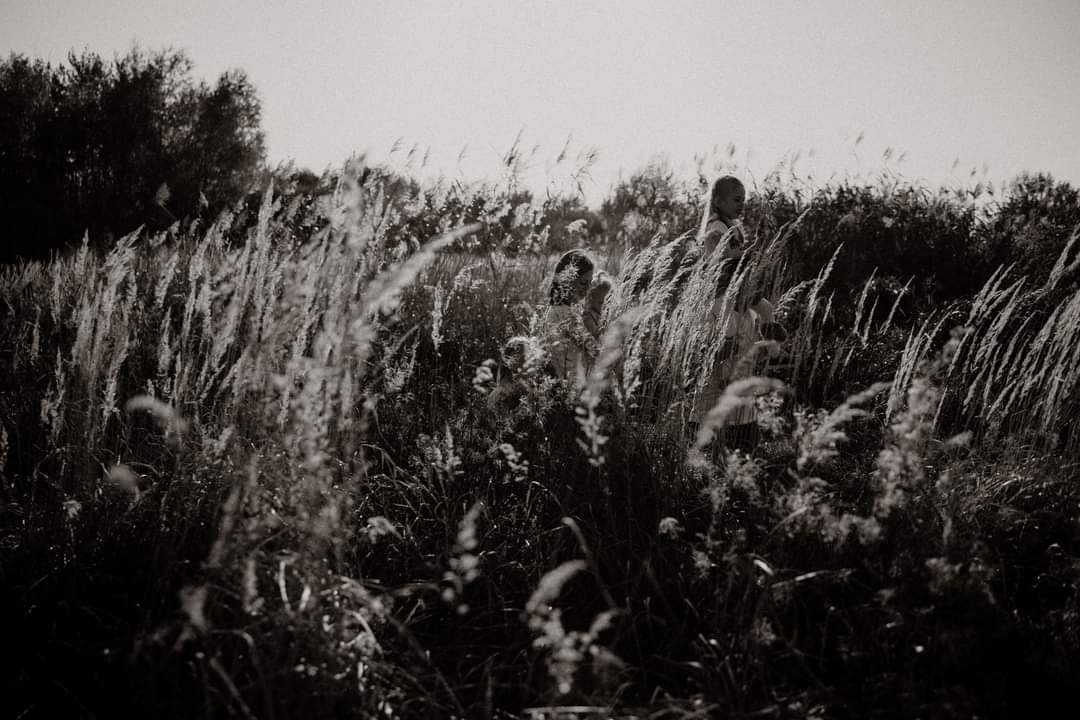 Czarno-białe zdjęcie dwóch dziewczynek stojących w wysokiej trawie na tle drzew, z silnym kontrastem i ziarnistością.