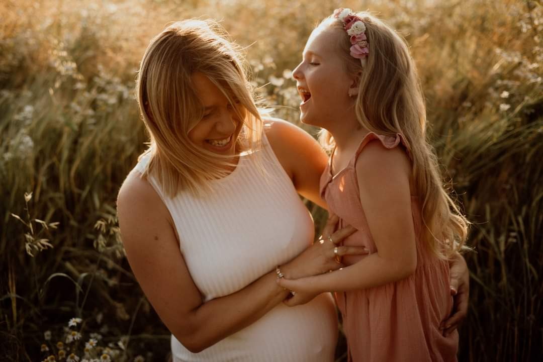 Ciężarna kobieta w białej sukience i dziewczynka w różowej sukience śmieją się na łące w promieniach zachodzącego słońca. Dziewczynka ma wianek na głowie.