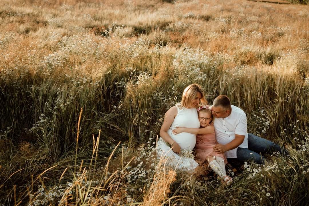 Ciężarna kobieta w białej sukience, mężczyzna w białej koszuli i dziewczynka w różowej sukience siedzą razem na łące z wysoką trawą i białymi kwiatami podczas słonecznej sesji.