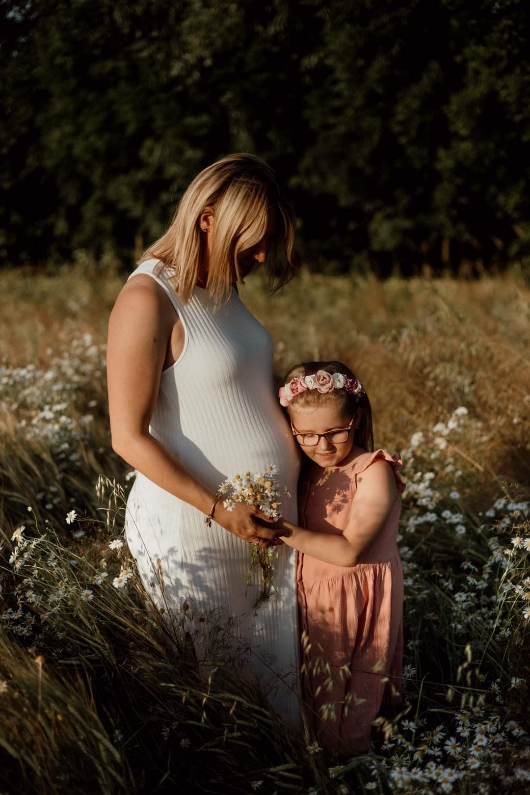 Kobieta w białej sukience ciążowej i dziewczynka w różowej sukience na łące z rumiankami, trzymają bukiet kwiatów, delikatne światło słoneczne.