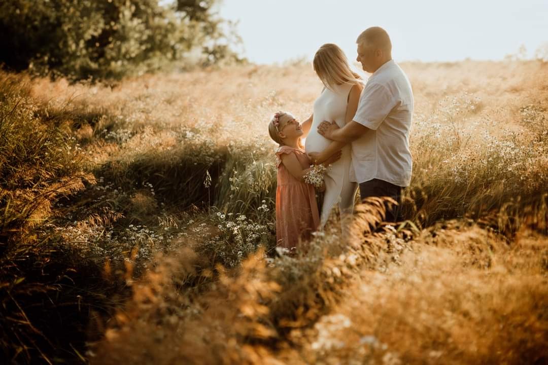 Przyszła mama w białej sukience, otoczona przez męża i córkę na łące w złotym świetle zachodzącego słońca, dziewczynka trzyma mały bukiecik białych kwiatów.