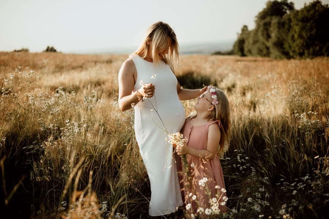 Ciężarna kobieta w białej sukience i dziewczynka z wiankiem na głowie stoją na łące pełnej polnych kwiatów, trzymając bukiety margaretek, w ciepłym świetle zachodzącego słońca.