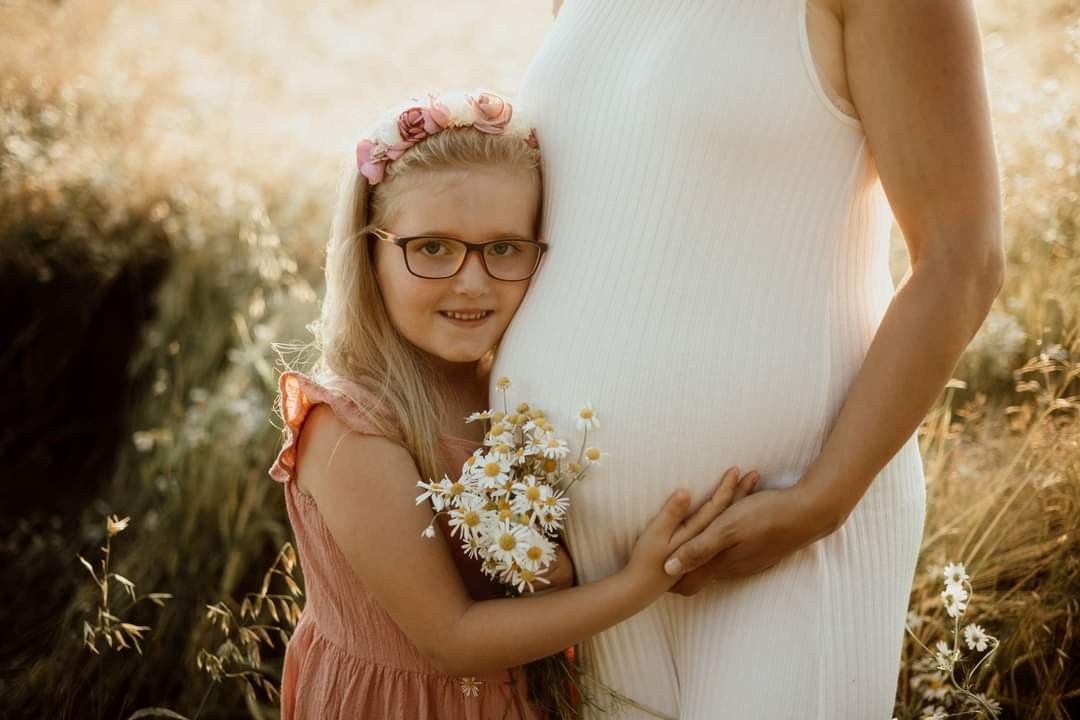 Dziewczynka w wianku z kwiatów przytula się do brzucha ciężarnej kobiety, trzymając bukiet białych stokrotek na tle łąki w złotym świetle.