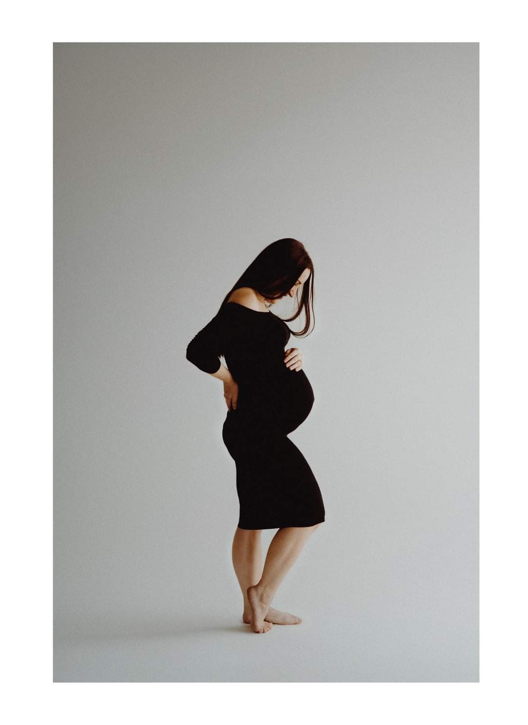 Kobieta w czarnej sukience, z rękami na brzuchu i biodrze, pozuje bokiem na neutralnym tle, eksponując ciążowy brzuszek, widoczne bose stopy.