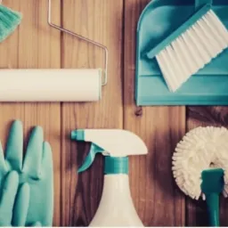 Zestaw narzędzi do sprzątania: miotła z szufelką, wałek do czyszczenia, szczotka do kurzu, rękawice gumowe i butelka ze spryskiwaczem na tle drewnianej ściany.