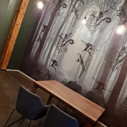 Jasne drewno stołu i szare krzesła w minimalistycznym biurze z tapetą przedstawiającą stylizowany las i ptaki oraz odsłoniętą drewnianą belką stropową.