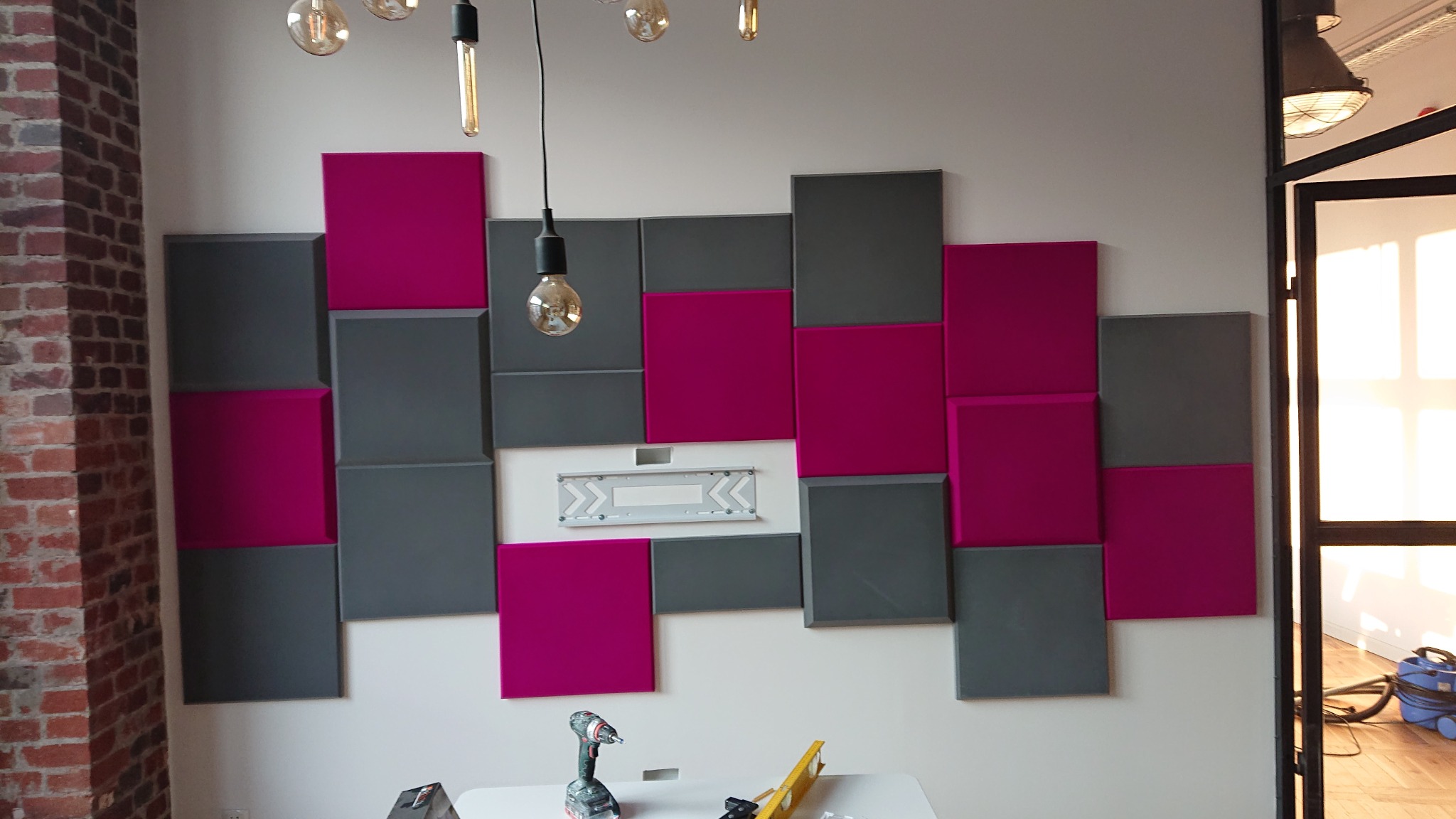 Montaż dekoracyjnych paneli ściennych w kolorze szarym i fuksji, widoczny uchwyt montażowy, wiertarka i poziomica na stole, ceglany filar po lewej stronie, surowe żarówki na suficie.