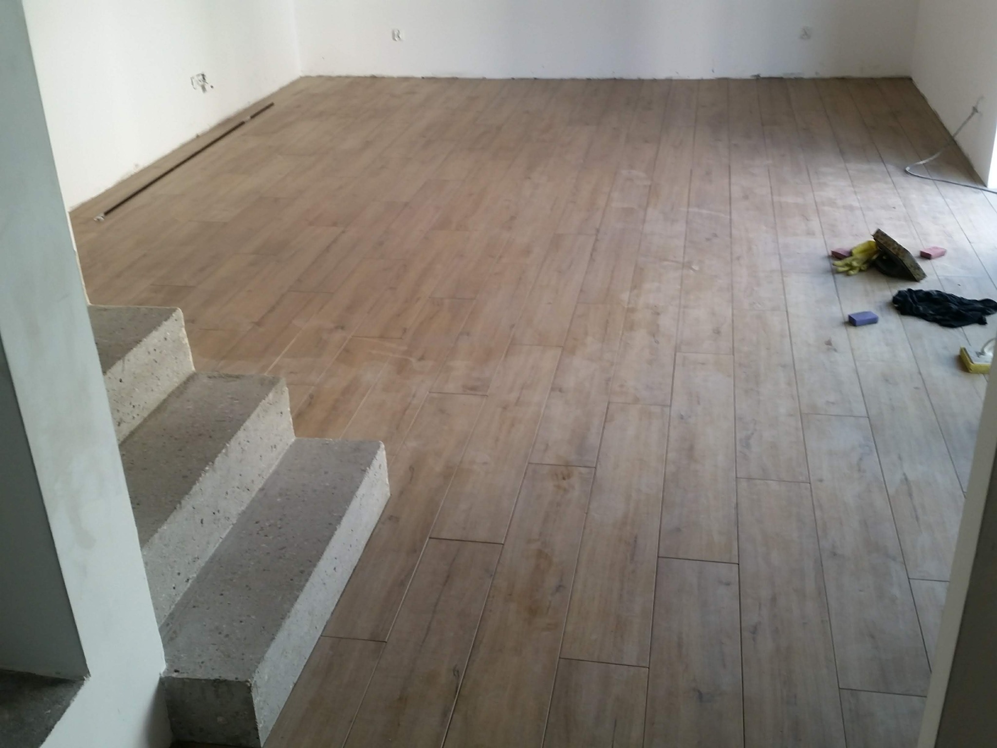 Ułożona podłoga z płytek imitujących drewno, widoczne betonowe schody i narzędzia glazurnicze w tle, minimalne ślady fugowania.