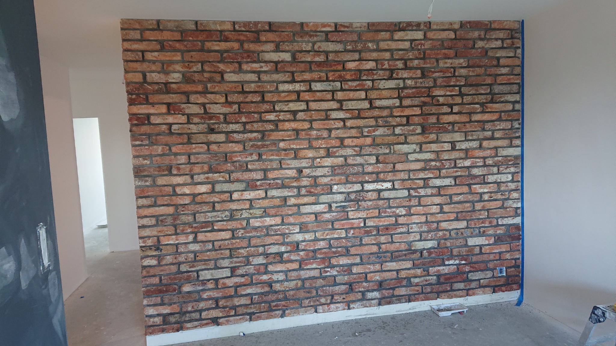 Ściana z cegły dekoracyjnej w trakcie montażu, zabezpieczona taśmą malarską, widoczne niedokończone wykończenia i akcesoria malarskie.