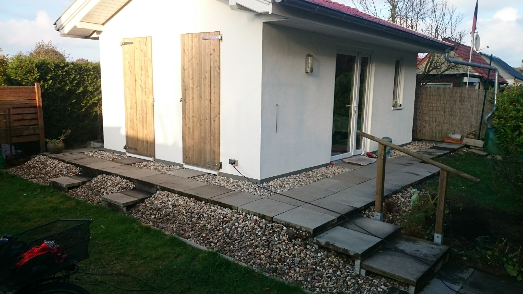 Biały budynek gospodarczy z drewnianymi drzwiami, wykończony ścieżką z płyt betonowych i kamieni ozdobnych, widok z zewnątrz.