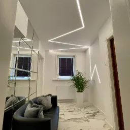 Nowoczesny korytarz z geometrycznym oświetleniem LED na suficie, lustrzaną ścianą, czarną sofą z szarymi poduszkami, marmurową podłogą i rośliną w białej donicy.