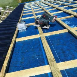 Montaż drewnianej konstrukcji dachu na niebieskiej membranie hydroizolacyjnej, widoczna czarna blacha dachowa, wkrętarka, pojemnik z wkrętami oraz pilarka tarczowa Bosch.