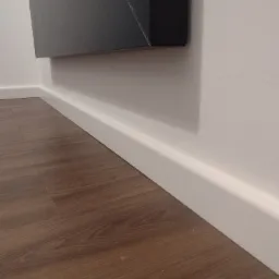 Ukośne ujęcie fragmentu ściany z zamontowaną białą listwą przypodłogową i panelami podłogowymi w odcieniach brązu, z narożnikiem czarnej dekoracji ściennej w lewym górnym rogu.