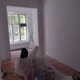 Pokój po świeżym malowaniu ścian na biało, zabezpieczone folią meble, widoczna drabina oparta o ścianę przy oknie i drewniana podłoga.