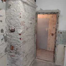 Ceglana ściana w trakcie prac przygotowawczych, pokryta warstwą szarego tynku, z widocznymi fragmentami cegieł i instalacją rurową przy suficie, otwór drzwiowy z widoczną ścianą w tle.