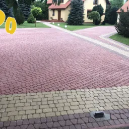 Czerwona kostka brukowa ułożona w jodełkę na podjeździe, z widoczną studzienką kanalizacyjną i otwartą, czarną bramą wjazdową, prowadząca do domu z ogrodem.