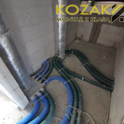 Bartosz Kozak Montaż z klasą - Rozprowadzenie przewodów wentylacyjnych i instalacji wodno-kanalizacyjnej w surowym wnętrzu, widoczne rury w izolacji oraz niebieskie i zielone przewody karbowane. W tle logo firmy.