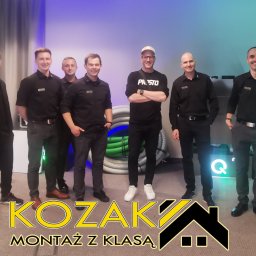 Bartosz Kozak Montaż z klasą - Zespół monterów w czarnych uniformach pozuje na tle zwiniętych rur wentylacyjnych i szarych rur kanalizacyjnych, widoczne logo firmy 'KOZAK Montaż z Klasą'.