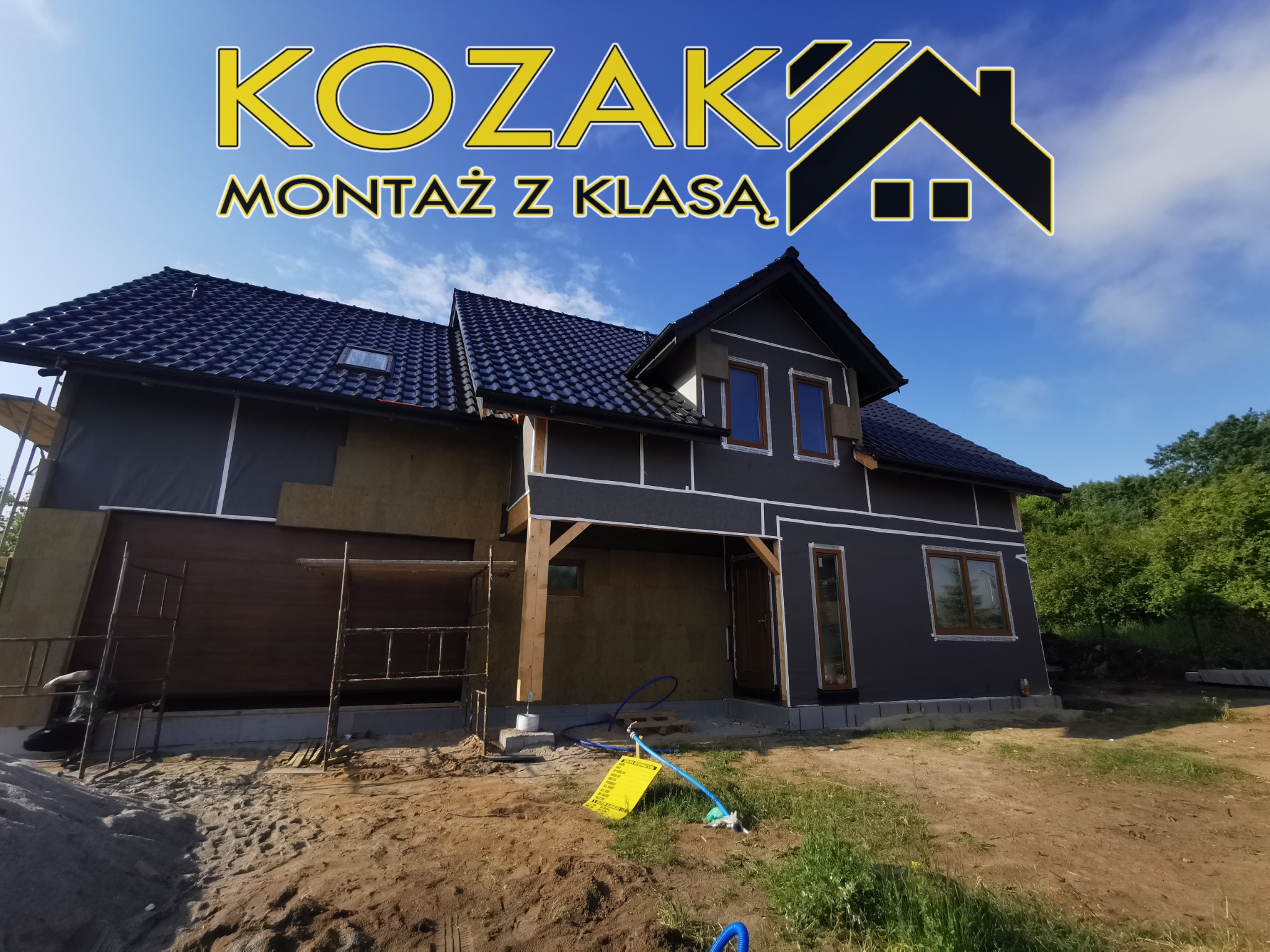 Dom w trakcie ocieplania, widoczne rusztowania, izolacja termiczna na ścianach i logo firmy 'KOZAK MONTAŻ Z KLASĄ' w górnej części kadru.