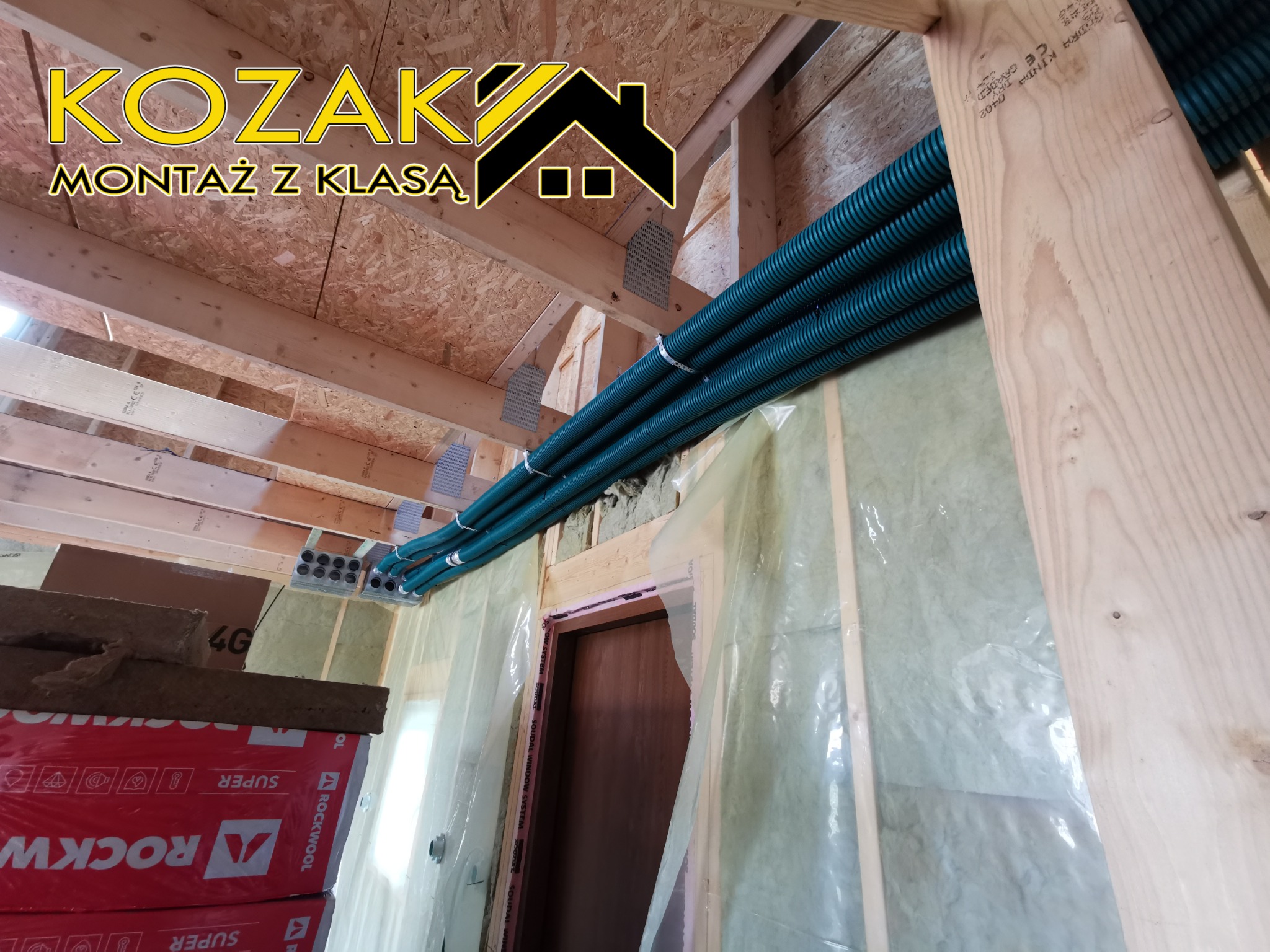 Wiązka zielonych, karbowanych rur wentylacyjnych zamontowana w surowym wnętrzu budynku w trakcie budowy, widoczne drewniane belki konstrukcyjne, izolacja z wełny mineralnej Rockwool i folia...