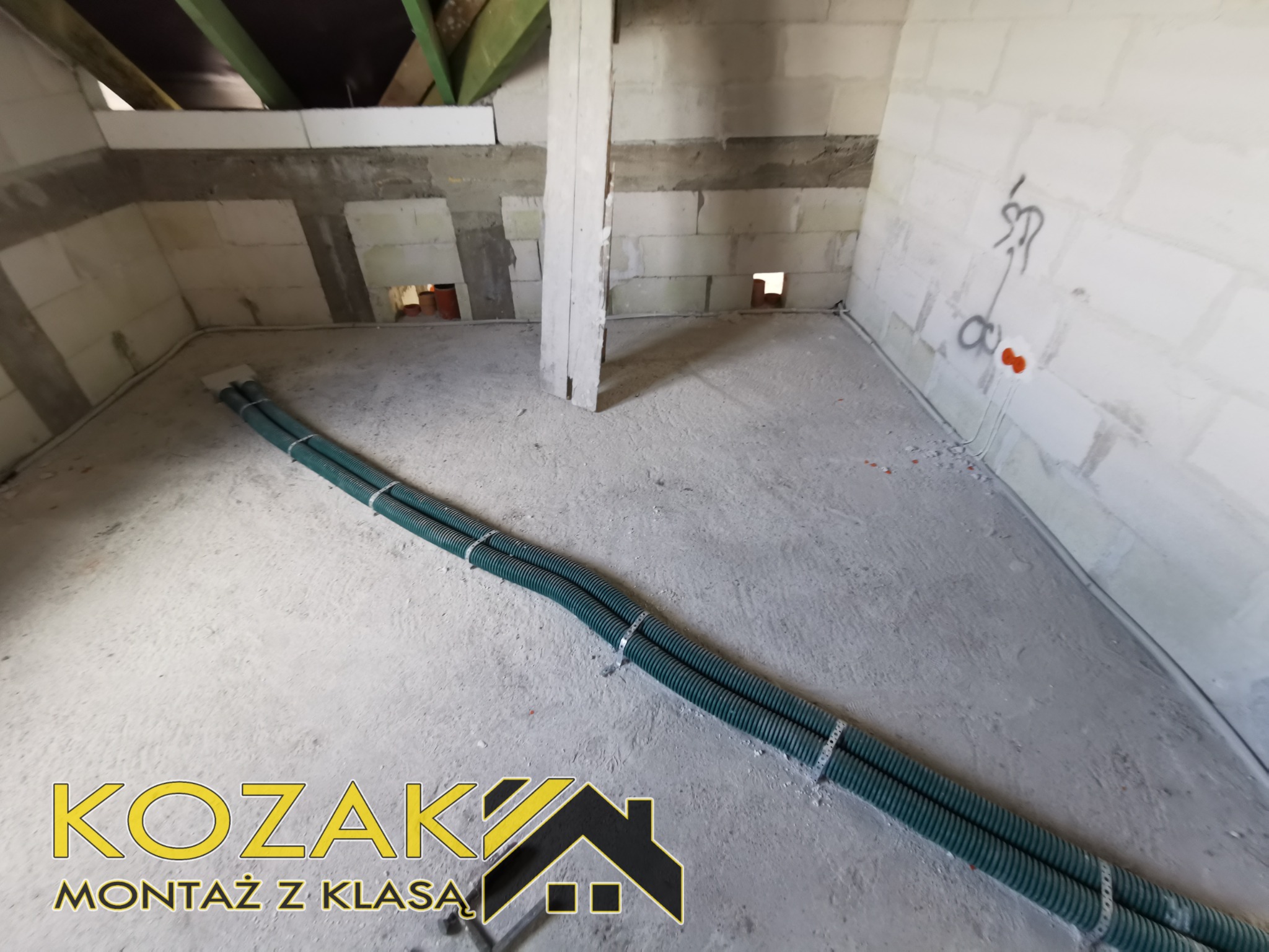 Dwa zielone karbowane przewody ułożone na szarym betonowym podłożu w surowym wnętrzu budynku w trakcie prac instalacyjnych. Na ścianach widoczne bloczki betonowe i otwory. W tle fragment...