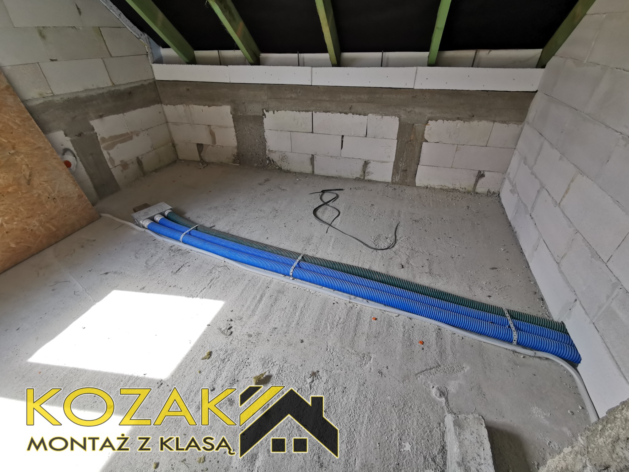 Niebieskie i zielone rury karbowane ułożone w korytku na surowej podłodze w trakcie prac instalacyjnych w budynku z pustaków, widoczny fragment instalacji elektrycznej i logo firmy.