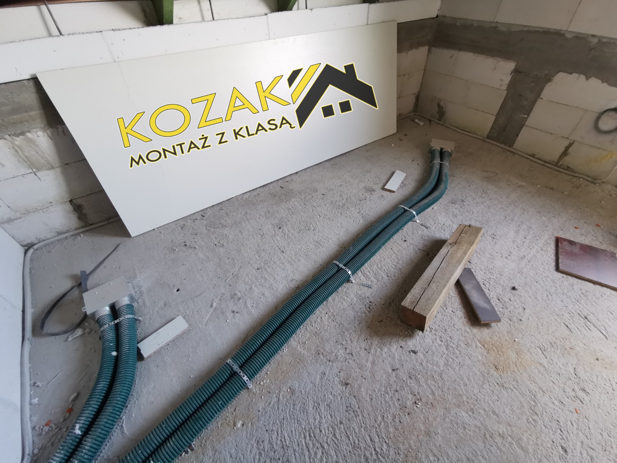 Zielone rury karbowane prowadzone po surowej podłodze, z logo firmy 'KOZAK MONTAŻ Z KLASĄ' na białej płycie w tle, widok z góry na niedokończone wnętrze.