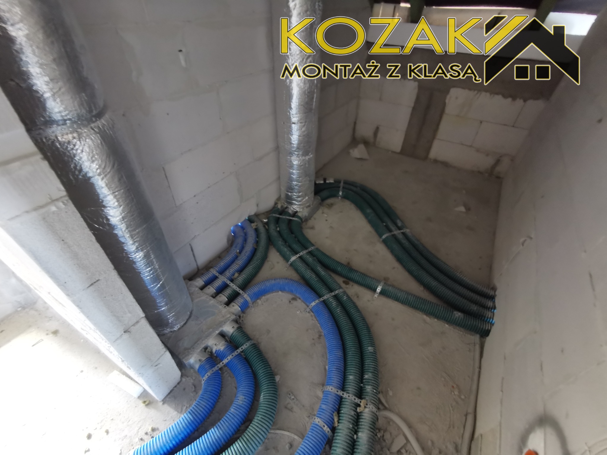 Rozprowadzenie przewodów wentylacyjnych i instalacji wodno-kanalizacyjnej w surowym wnętrzu, widoczne rury w izolacji oraz niebieskie i zielone przewody karbowane. W tle logo firmy.